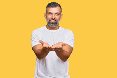Orta yaşlı, beyaz tişört giyen, elleri avuçlarında gülümseyen ve el hareketi yapan yakışıklı bir adam. Bekleme ve koruma 