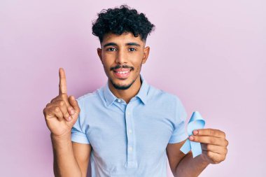 Genç Arap yakışıklı adam elinde mavi kurdele ile gülümsüyor bir fikirle ya da soru işareti yaparak mutlu bir yüzle, bir numara 