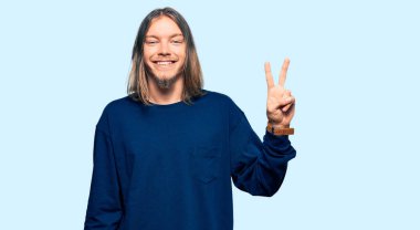 Uzun saçlı yakışıklı beyaz adam... günlük kışlık süveter giyiyor... ve parmaklarını iki numara ile gösteriyor... kendinden emin ve mutlu gülümserken.. 