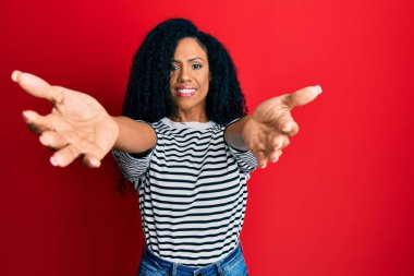 Orta yaşlı Afro-Amerikan kadın günlük giysiler içinde kameraya bakıp kucaklaşmak için gülümsüyor. Mutluluğu kucaklayan neşeli bir ifade.. 