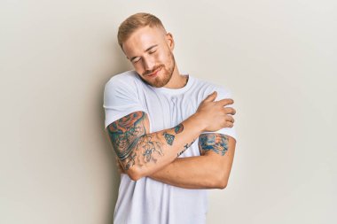 Beyaz günlük tişörtler giyen genç beyaz bir adam mutlu ve pozitif, kendinden emin bir şekilde kendine sarılıyor. Sevgi ve özgüven. 