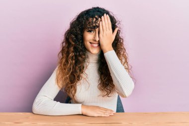 Latin asıllı genç bir kız gündelik giysiler içinde masada oturuyor bir gözünü eliyle kapatıyor, yüzünde kendine güvenen bir gülümseme ve sürpriz bir duygu var.. 