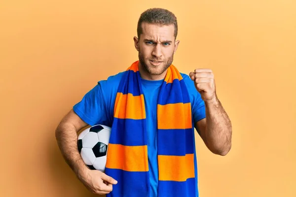 Young caucasian man football hooligan cheering game holding ball annoyed and frustrated shouting with anger, yelling crazy with anger and hand raised