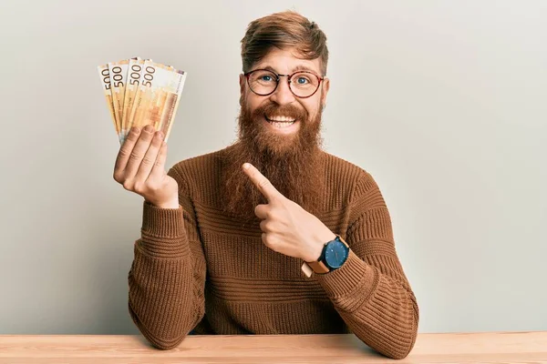 Young irish redhead man holding 500 norwegian krone banknotes sitting on the table smiling happy pointing with hand and finger