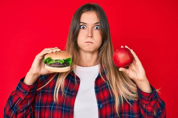 ハンバーガーと健康的な赤いリンゴの頬と面白い顔を比較する美しい白人女性 空気を吸い込み空気を吸い込み — ストック写真