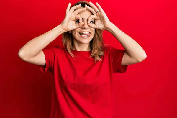 ヒスパニック系の若い女性は 双眼鏡の舌を突き出すようなOkジェスチャーを行うカジュアルな赤のTシャツを着て 目の指を見て クレイジーな表現 — ストック写真