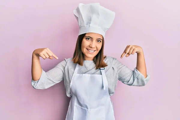 年轻美丽的女人穿着职业厨师的制服 头戴一顶帽子 面带微笑 用手指指着自己 自豪而快乐 — 图库照片