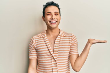 Makyaj yapan yakışıklı bir adam günlük tişört giyiyor neşeli bir şekilde gülümsüyor ve avuç içi kameraya bakıyor.. 