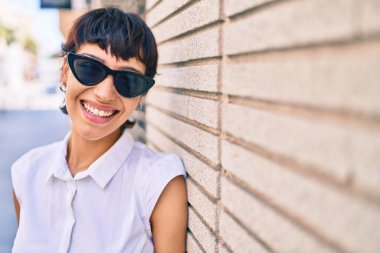 Kısa saçlı genç kadın dışarıda mutlu bir şekilde gülümsüyor güneş gözlüğü takıyor.