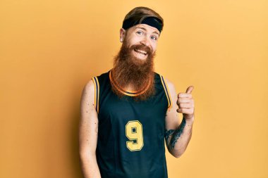 Uzun sakallı, basketbol forması giyen mutlu yüzlü, gülümseyen ve baş parmağıyla yan tarafı gösteren kızıl saçlı adam.. 