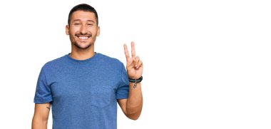 Dövmeleri olan yakışıklı bir adam. Günlük giysiler giyiyor. İki numarayı gösteriyor ve gülümsüyor.. 