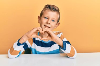 Sevimli beyaz çocuk, gündelik giysiler içinde masada oturmuş, aşkla gülümsüyor, elleriyle kalp sembolü yapıyor. romantik kavram. 