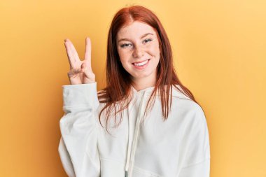 Genç kızıl saçlı kız rahat bir kazak giyer ve üç numaralı parmaklarıyla işaret ederken kendinden emin ve mutlu bir şekilde gülümser.. 