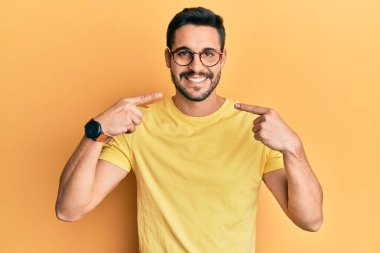 Gündelik kıyafetler giyen ve gözlüklü, neşeli gülümseyen, parmaklarıyla ve ağzıyla işaret eden İspanyol genç bir adam. diş sağlığı kavramı. 