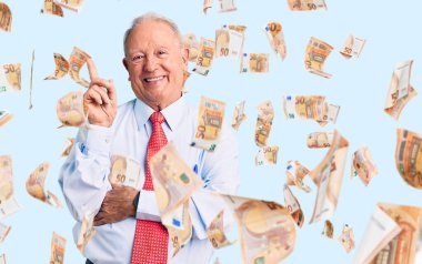 Zarif kravat ve gömlek giyen, yüzünde kocaman bir gülümseme olan, eli ve parmağı kameraya bakan, yakışıklı, gri saçlı bir adam..