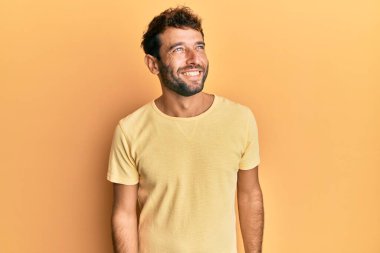 Sarı arka planda sarı tişört giyen sakallı yakışıklı bir adam yüzünde gülümseme, doğal bir ifade ile yan yana bakıyor. Kendinden emin gülen. 