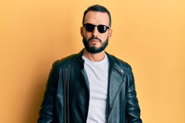 Sakallı, siyah deri ceketli ve yüzünde ciddi bir ifade olan güneş gözlüklü genç bir adam. Basit ve doğal bir şekilde kameraya bakıyorsun.. 