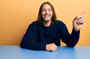 Yakışıklı, uzun saçlı, sıradan kıyafetler giyen, yüzünde kocaman bir gülümsemeyle masada oturan, eli ve parmağı kameraya bakan beyaz bir adam.. 