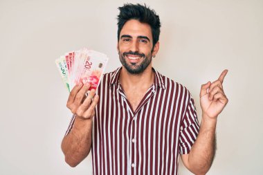 Sakallı yakışıklı İspanyol adam İsrail şekelleri tutuyor. Gülümsüyor. Elleri ve parmakları yana doğru uzanıyor. 
