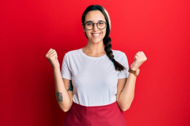 Profesyonel garson önlüğü giyen genç İspanyol kadın kollarını kaldırıp gözleri açık bir şekilde başarıyı kutluyor ve şaşırıyor. Kazanan konsept. 