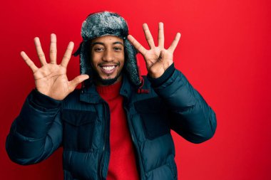 Sakallı, kışlık şapkalı ve paltolu genç Afrikalı Amerikalı dokuz numaralı parmaklarıyla kendini beğenmiş ve mutlu gülümserken kendini gösteriyor.. 