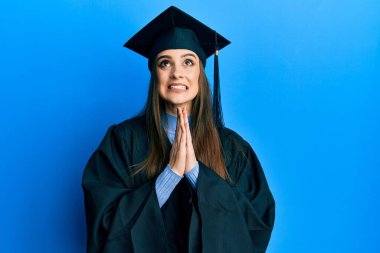 Güzel esmer bir kadın mezuniyet şapkası ve tören cübbesi takıyor. Elleriyle yalvarıyor ve dua ediyor. Yüzünde umut ifadesi var. Yalvarıyor. 