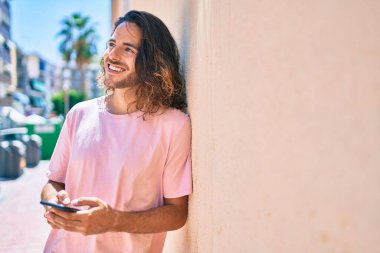 Duvara dayanmış akıllı telefon kullanan, mutlu gülümseyen genç İspanyol adam.