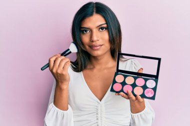 Makyaj fırçası ve allık paleti tutan genç Latin transseksüel kadın yüzünde ciddi bir ifadeyle rahatlamış. Basit ve doğal bir şekilde kameraya bakıyorsun.. 