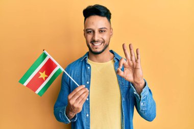 Surinam bayrağı tutan genç Arap adam parmaklarıyla işaret veriyor, gülümseyerek mükemmel bir sembol oluşturuyor. 