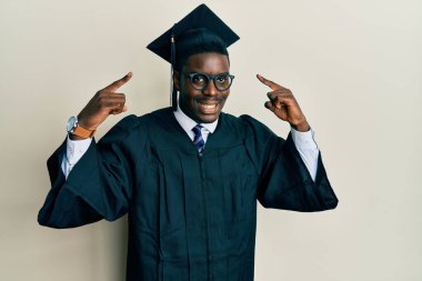 Mezuniyet şapkası ve tören cübbesi giyen yakışıklı siyah adam gülümseyerek iki eliyle başını işaret ediyor, harika bir fikir ya da düşünce, iyi bir hafıza. 