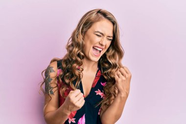 Gündelik giysiler giyen genç sarışın kız şaşkınlık ve şaşkınlık içinde kollarını kaldırarak ve gözlerini kapayarak başarıyı kutluyor. 