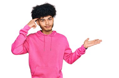 Afro saçlı, günlük pembe kazaklı, kafası karışmış ve avuç içi açık, fotokopi alanı gösteren ve alnını işaret eden Afro-Amerikan genç bir adam. Bunu bir düşün.. 