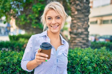 Mutlu gülümseyen ve içen genç sarışın kadın parktaki kahveyi alıp götürüyor..