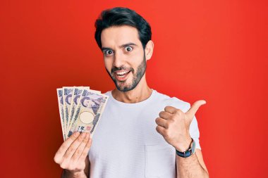 Japon yen banknotları tutan İspanyol genç adam başparmağıyla yan tarafı işaret ediyor, ağzı açık gülümsüyor. 