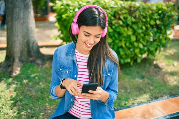 公園に座っているスマートフォンやヘッドフォンを使用して幸せな笑顔若い美しいヒスパニック系の女の子 — ストック写真