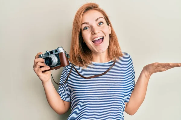 Young caucasian woman holding vintage camera celebrating achievement with happy smile and winner expression with raised hand