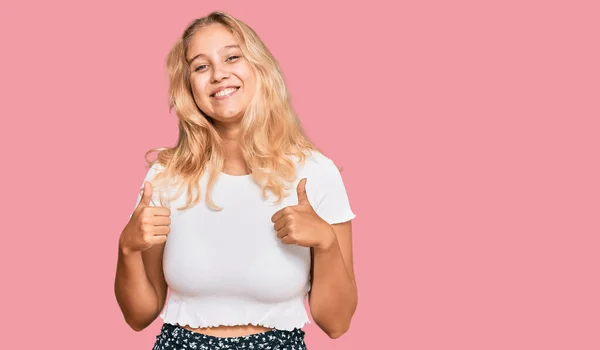 若いブロンドの女の子は 手で積極的なジェスチャーを行うカジュアルな白いTシャツの成功のサインを着て 親指を笑顔と幸せ 陽気な表情と勝者のジェスチャー — ストック写真