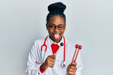 Genç Afrikalı Amerikalı kadın doktor üniforması giyiyor elinde çatal tutarak komik bir ifadeyle dilini dışarı çıkarıyor.. 