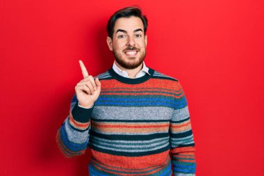 Sakallı, zarif yün kazaklı, yüzünde kocaman bir gülümseme olan, eli ve parmağı kameraya bakan yakışıklı beyaz bir adam.. 