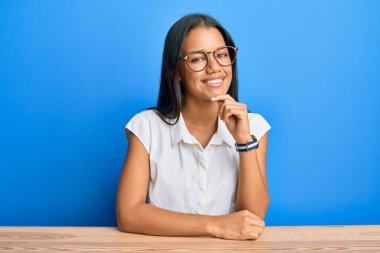 Güzel İspanyol kadın, gündelik giysiler içinde masaya oturmuş kameraya kendinden emin bir şekilde bakıyor kollarını kavuşturmuş ve çenesini kaldırmış gülümsüyor. Olumlu düşünmek. 