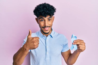 Genç Arap yakışıklı adam mavi kurdele tutuyor mutlu ve pozitif gülümsüyor, başparmağıyla mükemmel ve onay işareti yapıyor. 