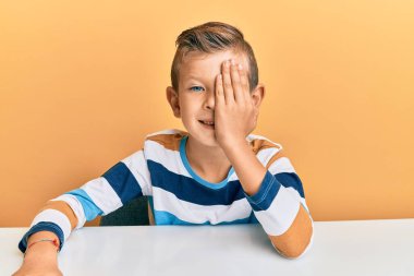 Beyaz elbiseli sevimli bir çocuk masanın üzerine oturmuş bir gözünü eliyle kapatıyor, yüzünde kendine güvenen bir gülümseme ve sürpriz bir duygu var.. 