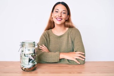 Masada oturan genç esmer kadın, elinde bir kavanoz dolusu dolarla kollarını kavuşturup kameraya bakarken gülümseyen mutlu yüzle. pozitif kişi. 