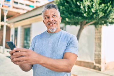 Orta yaşlı, ispanyol, kır saçlı, mutlu gülümseyen adam şehirde akıllı telefon kullanıyor..