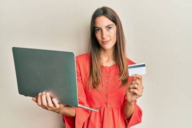 Dizüstü bilgisayar ve kredi kartı tutan genç ve güzel bir kadın yüzünde ciddi bir ifadeyle rahatlamış. Basit ve doğal bir şekilde kameraya bakıyorsun.. 