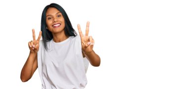 Gündelik giysiler giymiş genç Afro-Amerikan kadın kameraya bakıp zafer işareti yapıyor. İki numara.. 