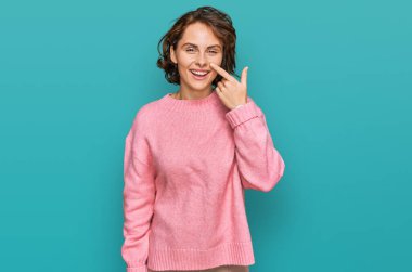 Latin asıllı genç bir kadın, gündelik giysiler giyip el ve burnu işaret ediyor, neşeli bir şekilde gülümsüyor. güzellik konsepti 