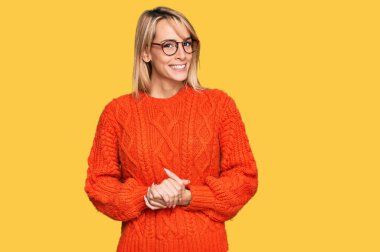 Güzel sarışın kadın, günlük kıyafetler ve gözlük takıyor ve ellerini kavuşturup gülümsüyor. Rahatlamış ve neşeli. Başarı ve iyimserlik 