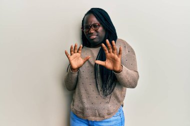 Saçları örgülü ve gözlüklü genç siyah kadın tiksindirici bir ifade takınıyor ve tiksinti tepki verdiği için tiksindirici ve korkunç bir ifade takınıyor. Eller havada. 