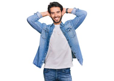 Genç İspanyol bir adam günlük kıyafetler giyiyor rahatlıyor ve esniyor, kolları ve elleri başının arkasında mutlu bir şekilde gülümsüyor. 